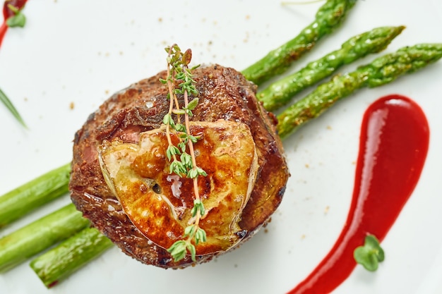 Portion gegrilltes Mignon-Steak mit Gänseleber und Spargel, Beerensauce in einem weißen Teller. Auf grauer Oberfläche isoliert.