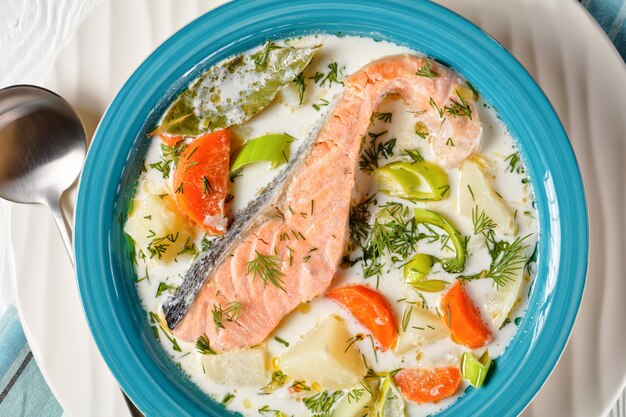 Portion Lachsfischsuppe mit Sahne, Kartoffeln, Karotten, Lauch und Dill in einer blauen Schüssel auf einem weißen Holztisch, finnische Küche, klassisches Gericht, horizontale Ansicht von oben