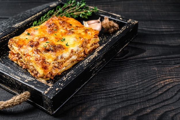 Portion Lasagne mit Rinderhackfleisch und Tomaten-Bolognese-Sauce in einem Holztablett. Schwarzer hölzerner Hintergrund. Ansicht von oben. Platz kopieren.