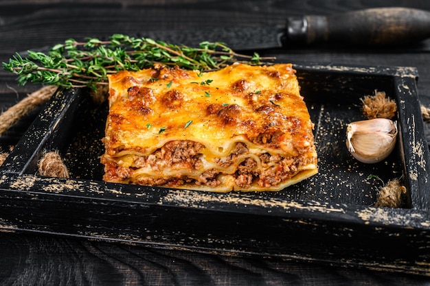 Portion Lasagne mit Rinderhackfleisch und Tomaten-Bolognese-Sauce in einem Holztablett. Schwarzer hölzerner Hintergrund. Ansicht von oben.