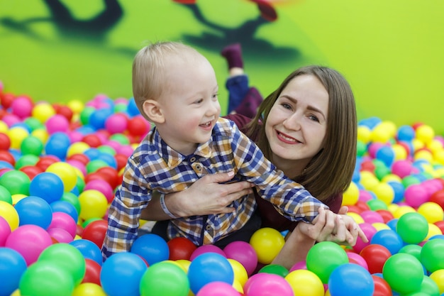 Porträt, das ein lächelnder junge spielt, wenn er mitte mit mutter spielt. lustiger junge im pool mit mehrfarbigen bällen. mutter und sohn haben spaß zusammen im spielzimmer hautnah. glückliche kindheit. familienwochenende.