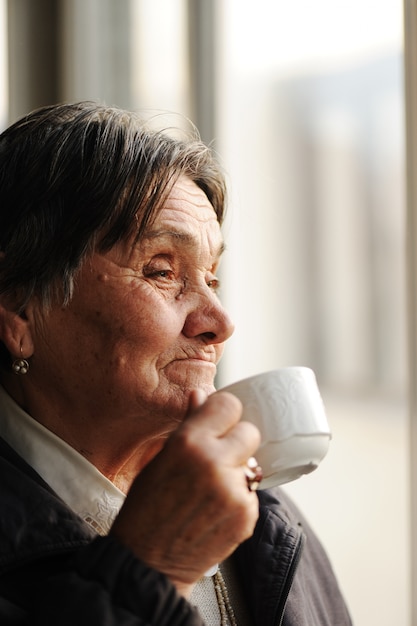 Porträt der älteren Frau schauend durch Fenster und trinkenden Kaffee