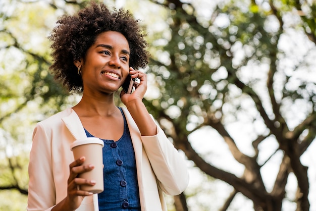 Porträt der Afro-Geschäftsfrau, die am Telefon spricht und eine Tasse Kaffee hält, während sie draußen im Park steht. Geschäftskonzept.