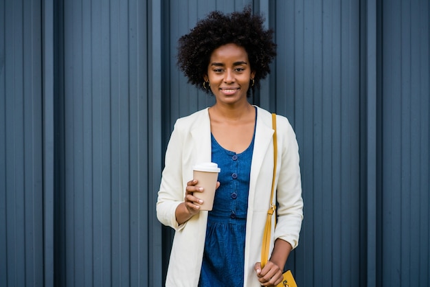Porträt der Afro-Geschäftsfrau, die eine Tasse Kaffee hält, während draußen auf der Straße steht. Geschäfts- und Stadtkonzept.