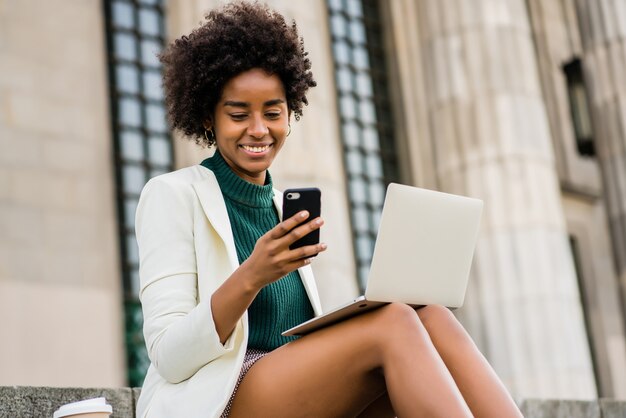 Porträt der Afro-Geschäftsfrau, die ihr Handy und Laptop beim Sitzen auf Treppen im Freien verwendet