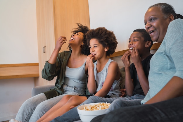 Porträt der afroamerikanischen Großmutter, der Mutter und der Kinder, die einen Film sehen und Popcorn essen, während sie zu Hause auf dem Sofa sitzen. Familien- und Lifestyle-Konzept.