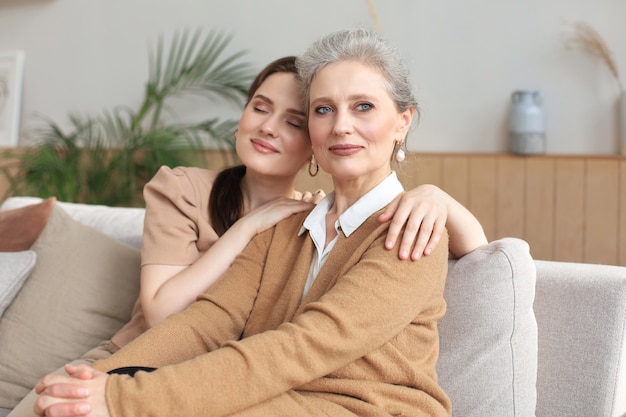 Porträt der alten Mutter und der reifen Tochter, die sich zu Hause umarmen. Glückliche ältere Mutter und erwachsene Tochter, die sich mit Liebe auf dem Sofa umarmen.