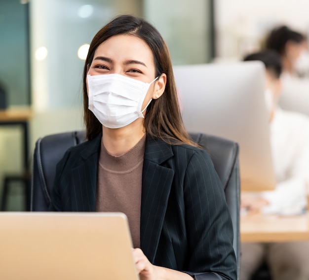 Porträt der asiatischen Büroangestellten Geschäftsfrau tragen schützende Gesichtsmaskenarbeit in neuem normalem Büro mit interracialem Kollegen im Hintergrund als soziale Distanzpraxis verhindern Coronavirus COVID-19.