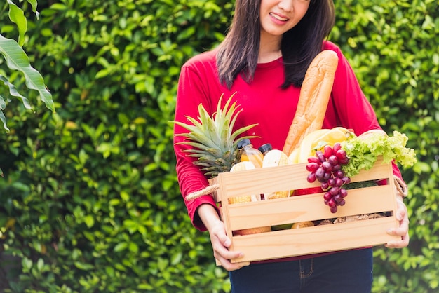 Porträt der asiatischen schönen jungen Bäuerin, die sie lächelt und frisches rohes Gemüse Obst in einer Holzkiste in ihren Händen auf grünem Blatthintergrund hält