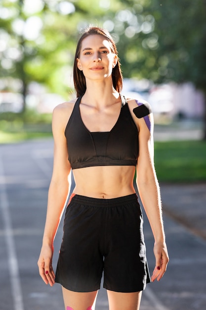 Porträt der atemberaubenden muskulösen Brunettefrau, die schwarzes Sportoutfit trägt, das draußen aufwirft. Junger Athlet, der im Stadion mit den Händen auf der Taille steht, bunte Behandlung am Körper, Kinesiotaping.