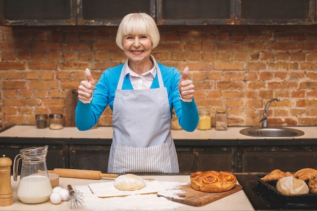 Porträt der attraktiven älteren älteren Frau kocht auf Küche. Großmutter macht leckeres Backen. Daumen hoch.