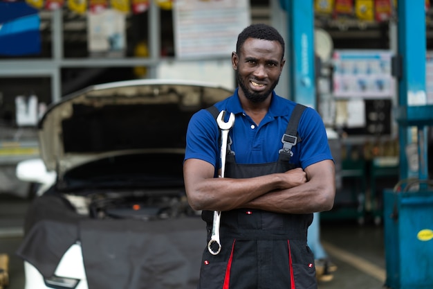 Porträt der attraktiven afrikanischen Mannarmkreuzblickkamera und der großen Schlüsselwerkzeuge in der Hand. Sachkundiger Mechaniker, der in einer Autowerkstatt arbeitet.