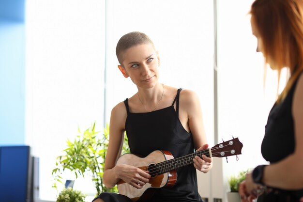 Porträt der attraktiven jungen Frau, die auf brauner Ukulele spielt und Zeit mit Freund verbringt. Kurzhaarige Dame im schwarzen Kleid. Großer heller Raum. Künstler- und Musikkonzept