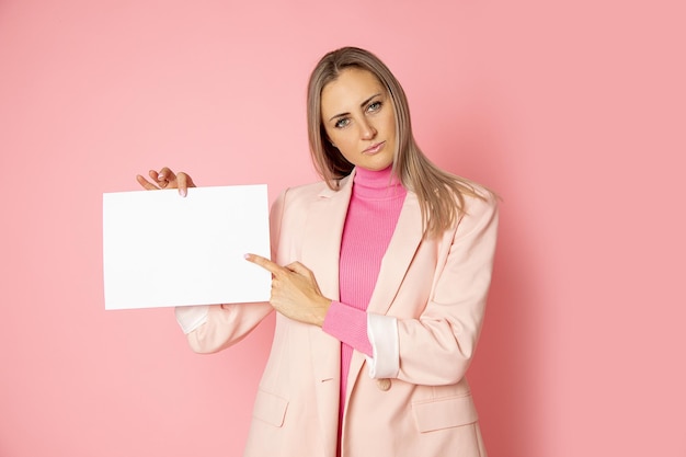 Porträt der attraktiven jungen Frau, die leeres Papier A4 für Zeichen auf rosa Hintergrund hält. Geschäftsfrau trägt rosa Jacke. Zeigt mit dem Finger auf den Kopierraum.