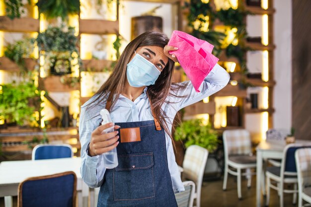 Porträt der attraktiven weiblichen Kellnerin, die Gesichtsmaske trägt, die eine Flasche mit Desinfektionsmittel und einem Lappen in einem Restaurant hält. Neue Normalität mit Coronavirus-Ausbruchshygiene-Restaurantkonzept.