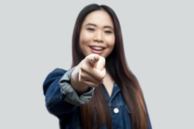 Porträt der aufgeregten asiatischen jungen Frau des schönen Brunette in der zufälligen blauen Denimjacke mit dem Make-up, das zahnig lächelt und auf die Kamera zeigt. Indoor-Studioaufnahme, isoliert auf hellgrauem Hintergrund.