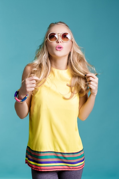 Porträt der bezaubernden schönen blonden Frau mit Sonnenbrille und gelbem Hemd auf blauem Hintergrund. Unbeschwerter Sommer.