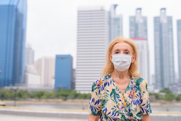 Porträt der blonden älteren Frau, die Maske zum Schutz vor Coronavirus-Ausbruch auf Ansicht der Stadt trägt