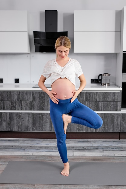 Porträt der blonden frau in der sportbekleidung in voller länge, die auf einem bein steht und den bauch streichelt und zu hause in der küche meditiert