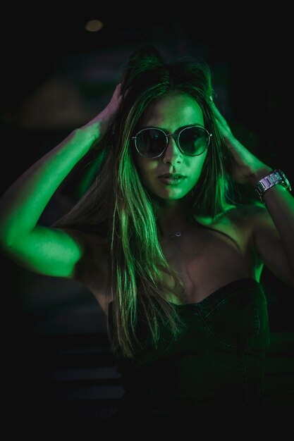 Porträt der brünetten kaukasischen Frau mit Sonnenbrille, die eine hellgrüne LED betrachtet. Nachtstadtfotografie