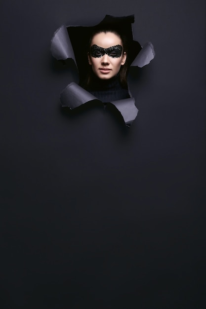 Porträt der charmanten eleganten brünetten Frau im schwarzen Rollkragenpullover und in der Paillettenmaske schaut durch ein zerrissenes Studio