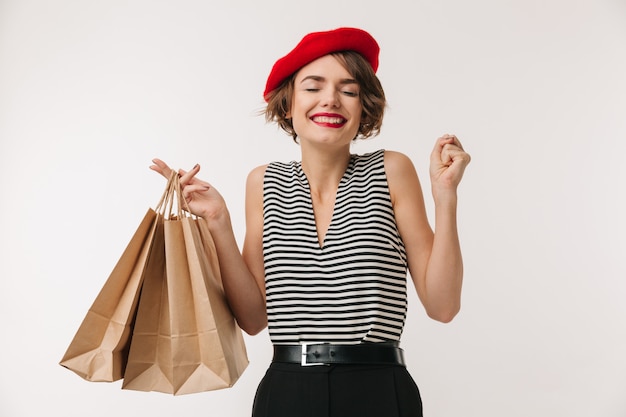 Porträt der eleganten Frau im gestreiften Hemd und im roten Baskenmütze lächelnd, während Einkaufstaschen getragen, lokalisiert über Weiß