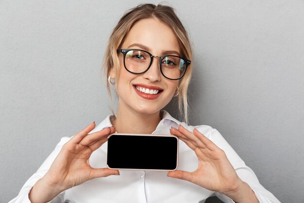 Porträt der entzückenden Geschäftsfrau, die eine Brille trägt, die lächelt und Smartphone im Büro hält, lokalisiert