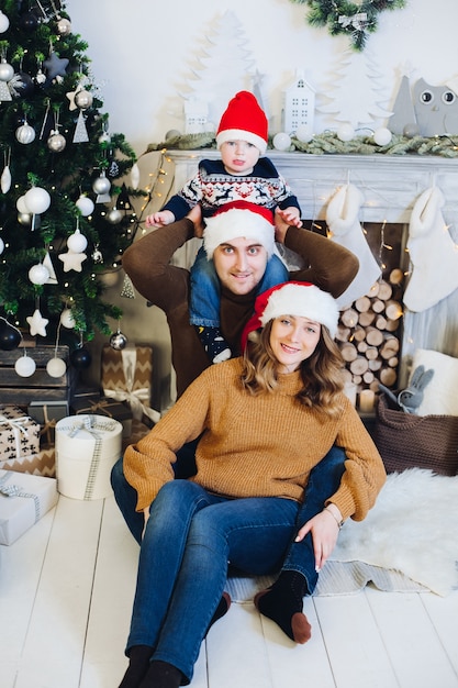 Porträt der entzückenden liebenden Familie mit dem Sohn auf den Schultern des Vaters, der vorne lächelnd am Kamin und am Weihnachtsbaum sitzt und alle Weihnachtsmützen trägt