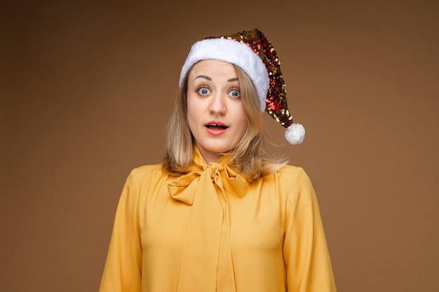 Porträt der erstaunten und schockierten blonden Frau, die gelbe elegante Bluse und funkelnde festliche Weihnachtsmütze trägt, die mit großen Augen schaut