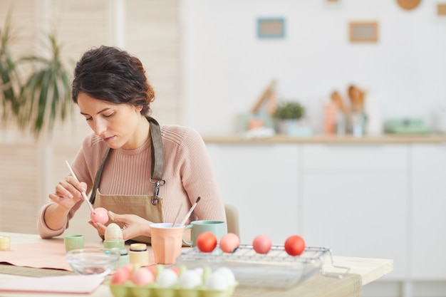 Porträt der erwachsenen Frau, die Eier in den Pastellfarben für Ostern malt, während am Tisch im gemütlichen Kücheninnenraum sitzen, Raum kopieren