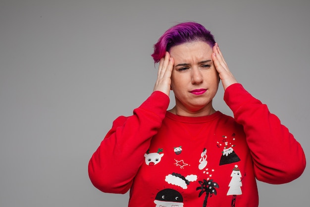 Porträt der erwachsenen Frau mit kurzen lila Haaren im roten Weihnachtspullover, der ihre Schläfen wegen Kopfschmerzen berührt. Speicherplatz kopieren