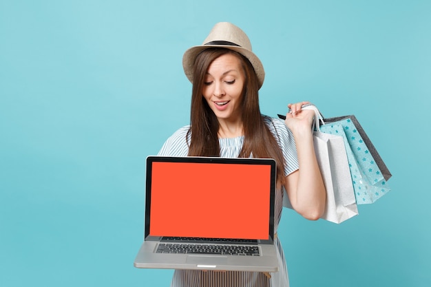 Porträt der Frau im Sommerkleid, Strohhut, der Pakettaschen mit Einkäufen nach dem Einkaufen hält, Laptop-PC mit leerem Bildschirm einzeln auf blauem Pastellhintergrund. Kopieren Sie Platz für Werbung.