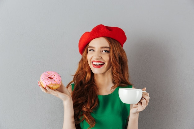 Porträt der freudigen eleganten Dame mit Ingwerhaar in der roten Baskenmütze beim Frühstück mit Tasse Kaffee und köstlichem Donut, lokalisiert über graue Wand