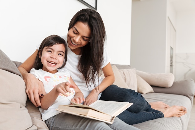 Porträt der freudigen Familienmutter und des freudigen Familienkindes, die Zeit zusammen verbringen, auf Sofa im Wohnzimmer sitzen und Buch lesen