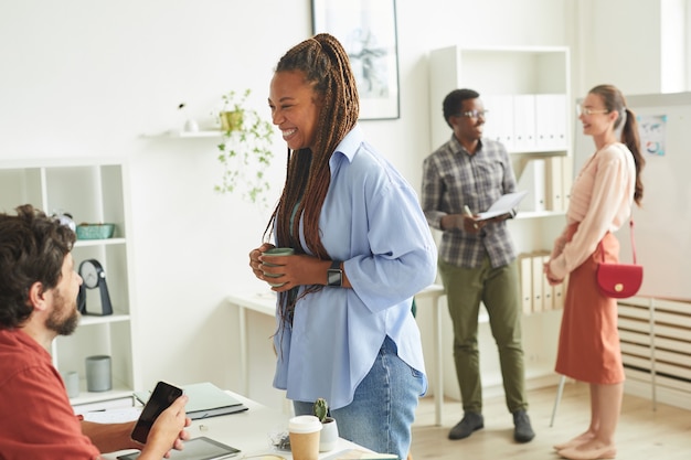 Porträt der fröhlichen afroamerikanischen Frau, die mit Kollege spricht, während sie im Büro am Tisch steht