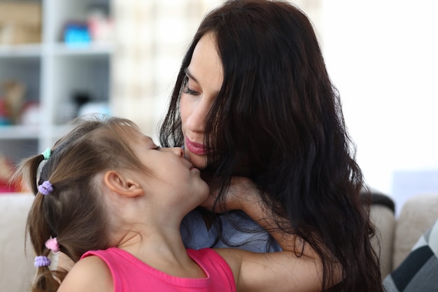 Porträt der fürsorglichen Mama und des fröhlichen Kindes, die genießen, lustige Zeit zusammen zu Hause zu verbringen. Schöne Mutter küsst glückliche Tochter. Konzept für Kindheit und Elternschaft