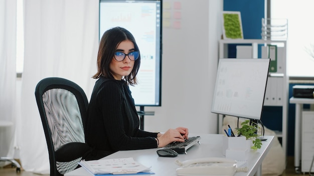 Porträt der Geschäftsfrau, die Computer für Diagrammdesign am Firmensitz verwendet. Ingenieur mit Unternehmensjob, der mit Technologie und Monitor für die Datenanalyse arbeitet und die Kamera betrachtet.