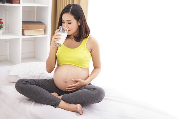 Porträt der gesunden schwangeren Frau, die ein Glas Milch trinkt, während auf Bett sitzend