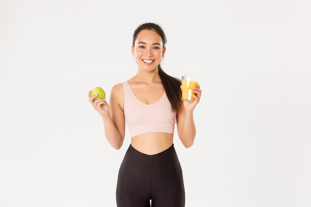 Porträt der gesunden und fit gut aussehenden asiatischen Sportlerin, die Orangensaft und Apfel hält, Morgenfrühstück vor dem Training, weißer Hintergrund.