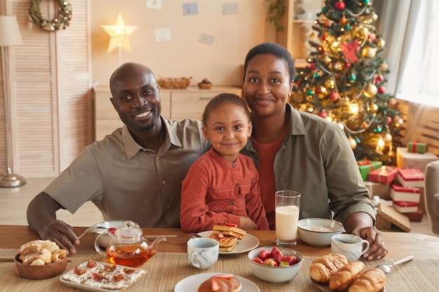 Porträt der glücklichen afroamerikanischen Familie, die Tee und Süßigkeiten genießt, während Weihnachten zu Hause im gemütlichen Innenraum feiert