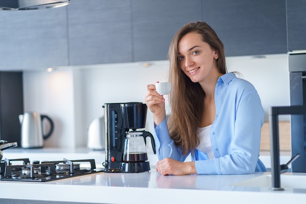 Porträt der glücklichen attraktiven Frau, die frischen aromatischen Kaffee trinkt, nachdem Kaffee mit Kaffeemaschine in der Küche zu Hause gebrüht wird. Kaffeemixer und Haushaltsküchengeräte für macht heiße Getränke