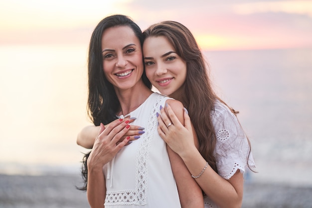Porträt der glücklichen attraktiven lächelnden umarmenden Tochter und Mutter im Sommer bei Sonnenuntergang am Meer