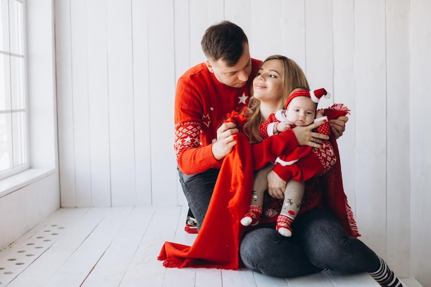 Porträt der glücklichen familie, mutter, vater und kleiner tochter in roter traditioneller weihnachtskleidung, die zeit zusammen in einem hellen holzzimmer in der nähe des fensterkopienraums verbringen