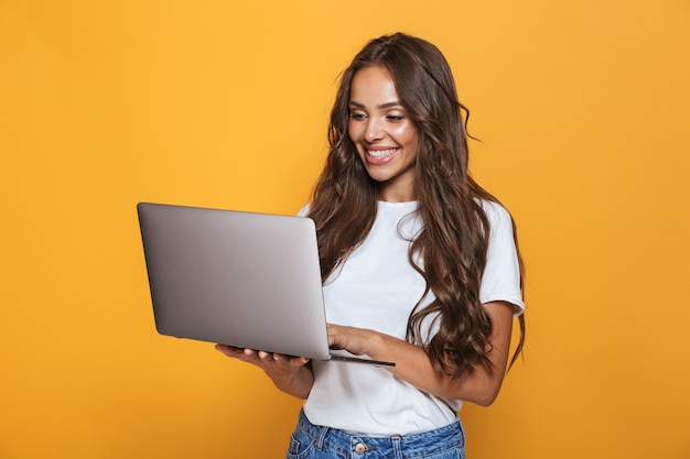 Porträt der glücklichen Frau 20s mit langen Haaren lächelnd und hält grauen Laptop, lokalisiert über gelbe Wand