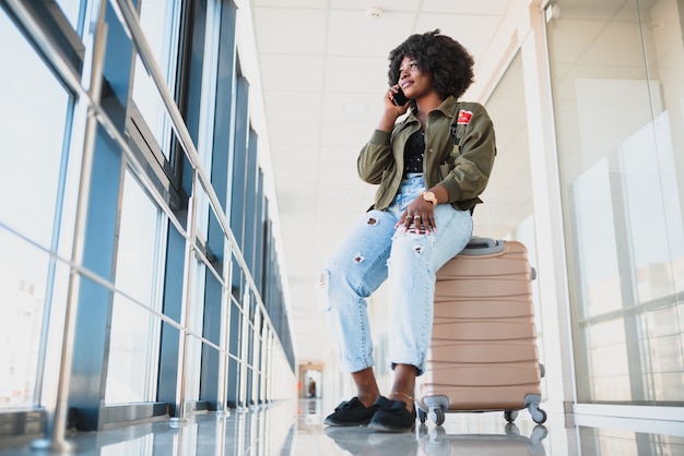 Porträt der glücklichen jungen Afroamerikanerfrau, die auf Koffer sitzt und mit Handy am Bahnhof spricht