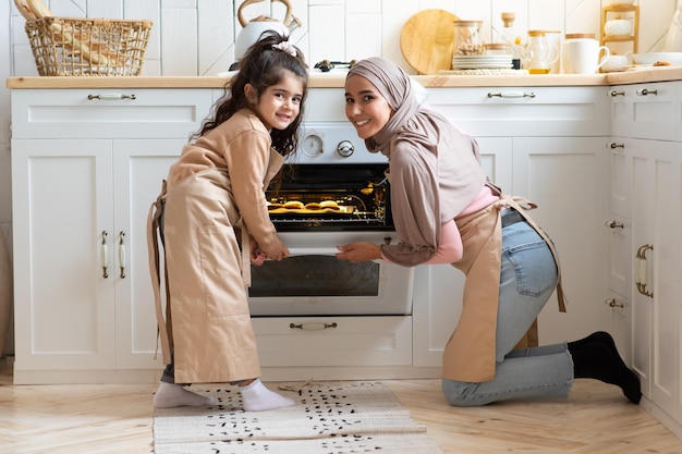 Porträt der glücklichen muslimischen Familienmutter und -tochter, die zusammen im Kücheninnenraum backen. Islamische Mutter und ihr Kind kochen hausgemachtes Gebäck, öffnen den Ofen mit frisch gebackenen Muffins im Inneren, freier Platz