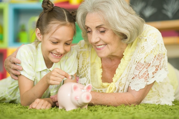 porträt der glücklichen schönen großmutter mit der enkelin, die münze in das sparschwein steckt