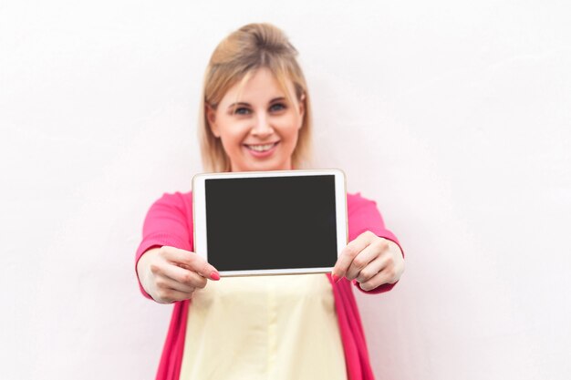 Porträt der glücklichen schönen jungen Geschäftsfrau in der rosa Bluse, die leeren Bildschirm der Tablette mit zahnigem Lächeln steht und hält und Kamera betrachtet. Innen, isoliert, Kopienraum, weißer Hintergrund