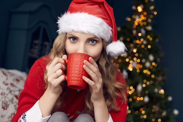 Porträt der herrlichen blonden Frau im roten Pullover und in der Weihnachtsmannmütze mit Tasse Kaffee, die im weihnachtlich geschmückten Innenraum aufwirft.