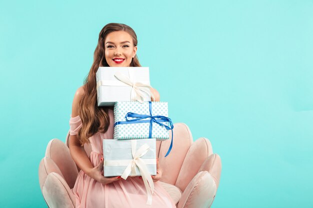 Foto porträt der herrlichen frau 20s mit langen braunen haaren, die viele geschenkboxen halten, während auf rosa sessel sitzen, lokalisiert über blauer wand